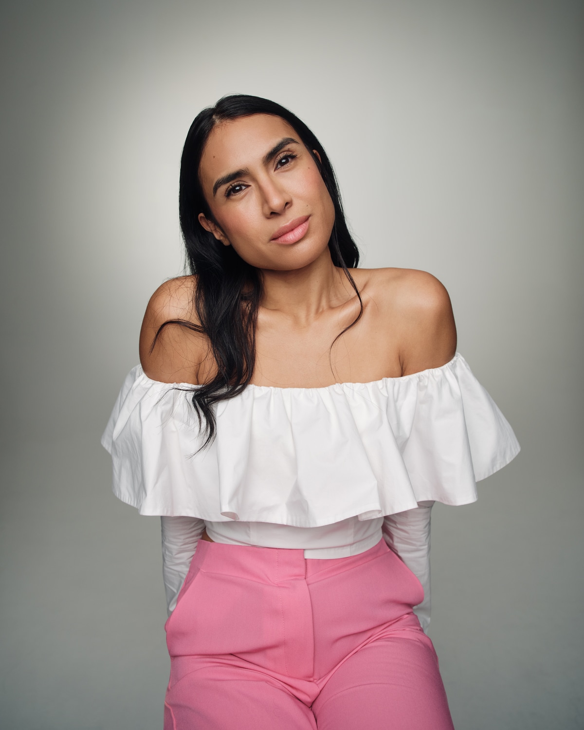 Yasmine Seifi in a white blouse and pink trousers against a grey background.