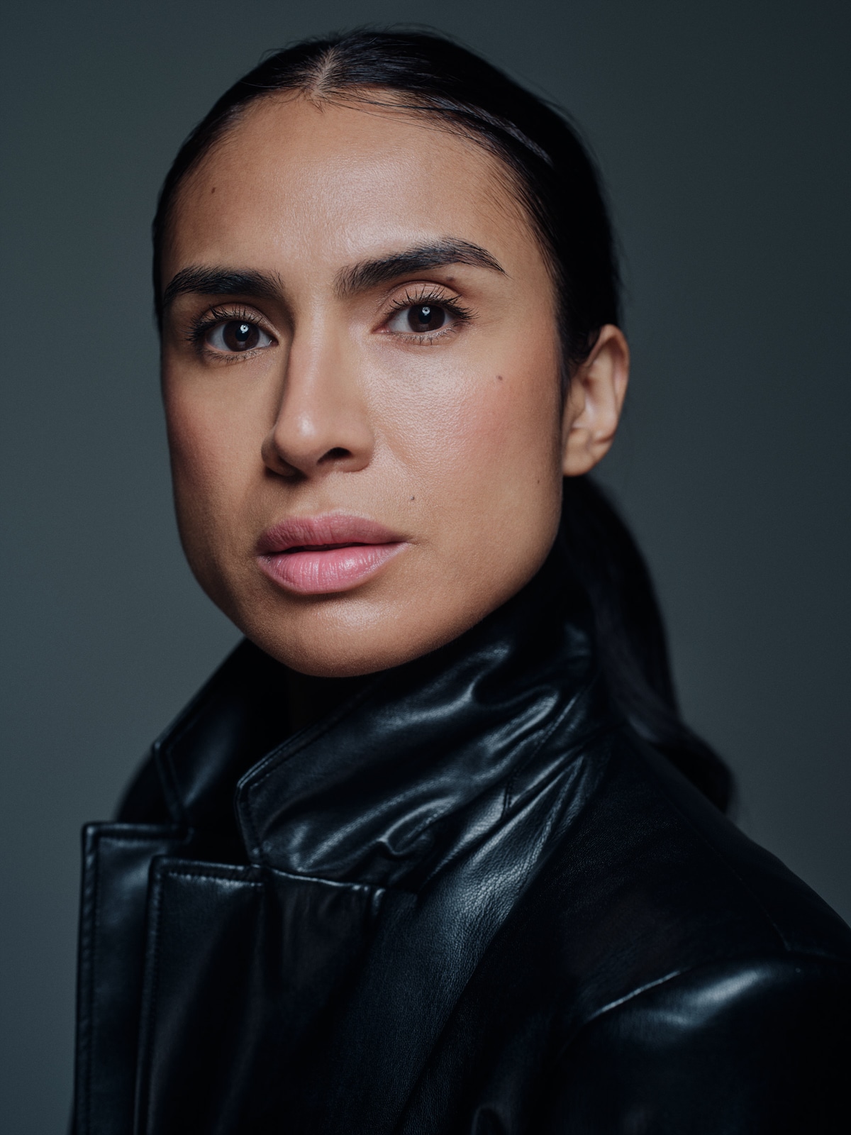 Close-up portrait of Yasmine Seifi with her hair tied back, wearing a black leather jacket against a dark grey background.