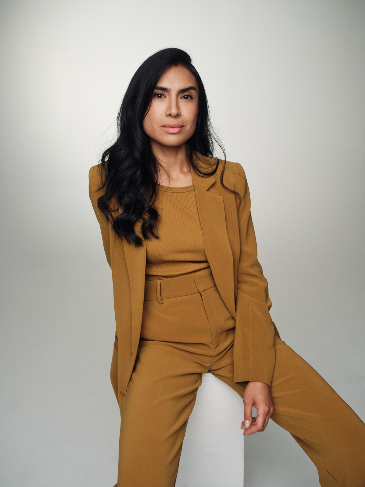 Yasmine Seifi seated in an ochre suit against a light grey studio background.
