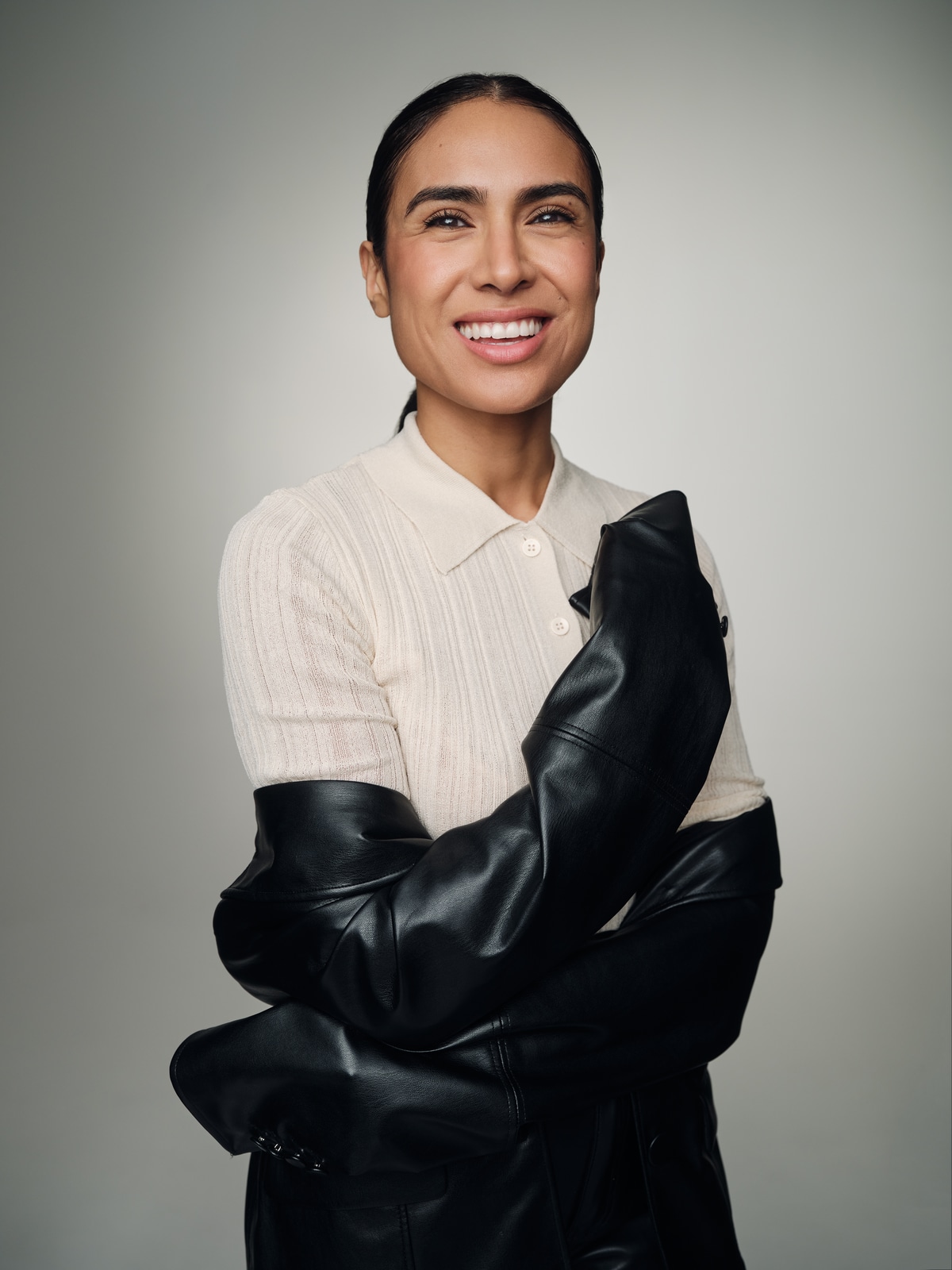 Yasmine Seifi smiling, wearing a cream knit shirt with a black leather jacket draped around her arms.